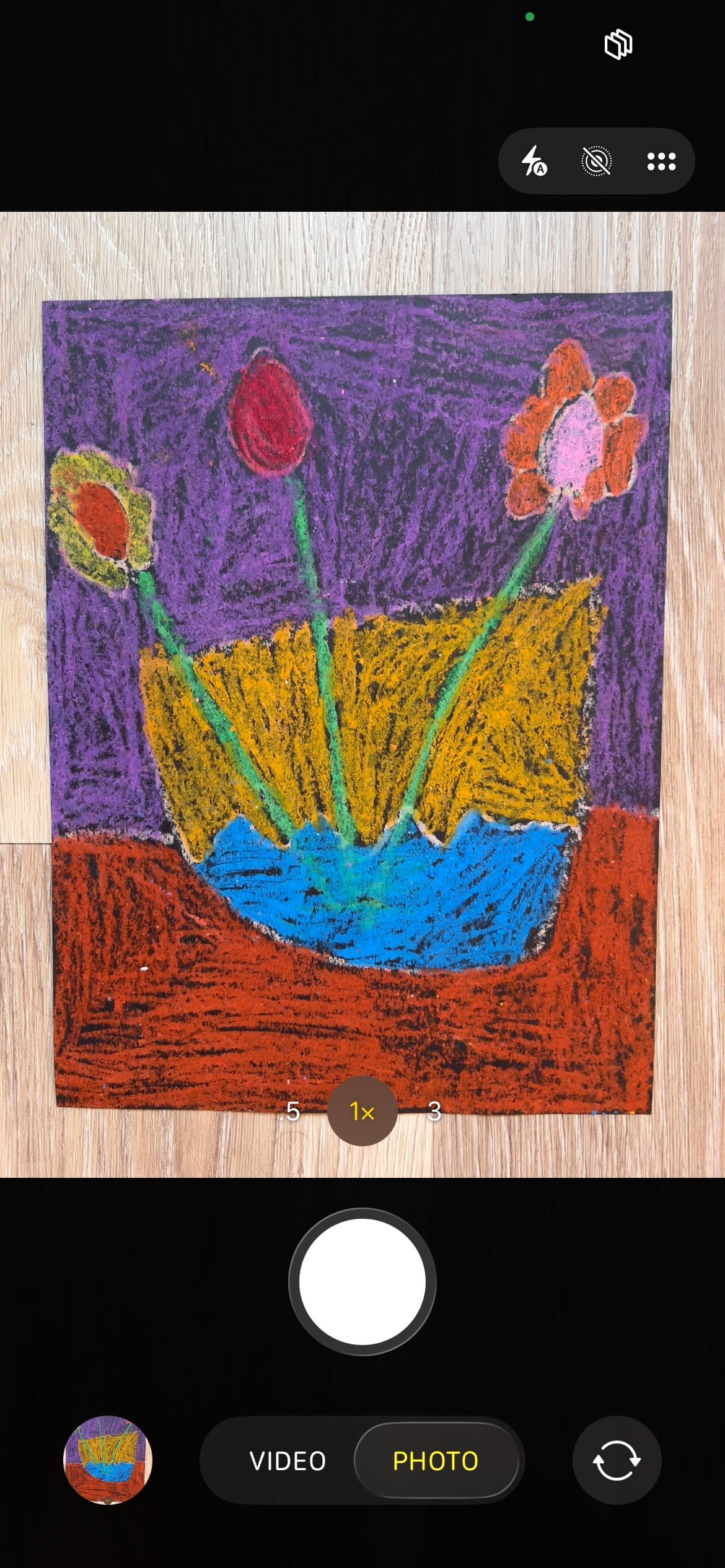 A phone camera framing a child's crayon drawing of flowers in a vase, ready to photograph