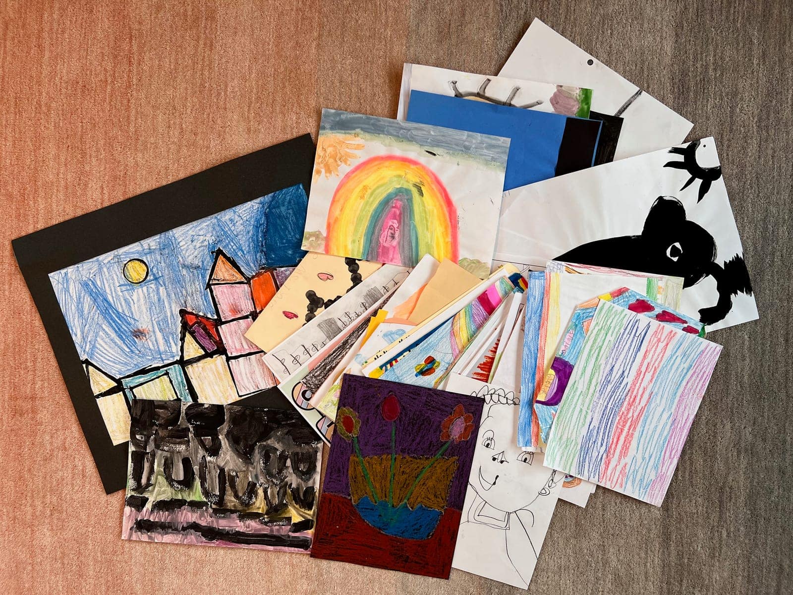 A pile of children's artwork on the floor, crayon drawings, paintings, and construction paper stacked and overlapping